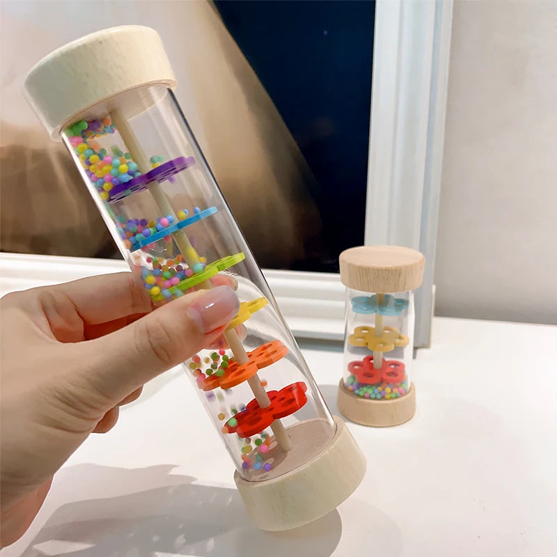 A hand holds a wooden rain sound tube musical instrument filled with colorful plastic beads and sticks.