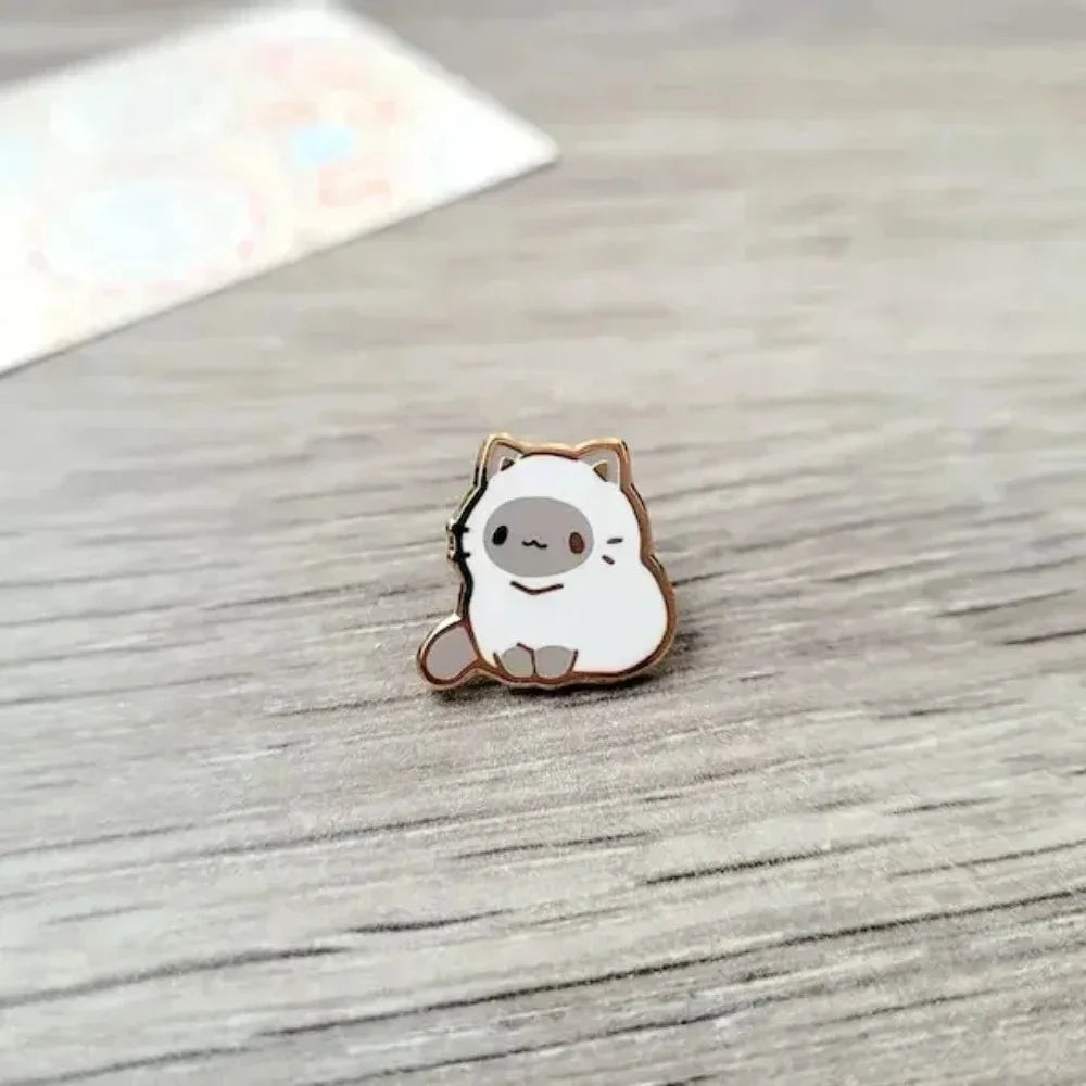 Un pequeño broche de esmalte redondo que representa la cara de un gato blanco lindo con acentos marrones.