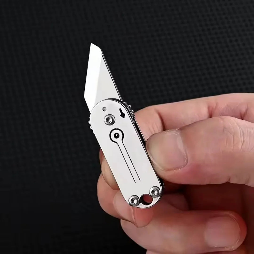A hand holds a compact stainless steel pocket knife with a visible blade and keychain mechanism.