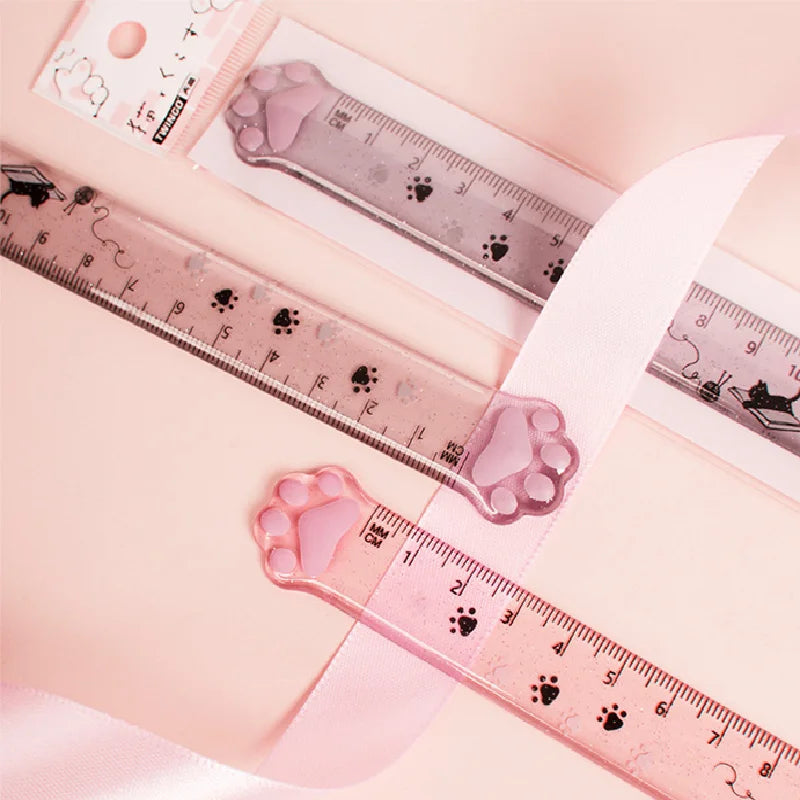 A plastic ruler marked with cat paw prints is shown next to a pink ribbon and a small product label.