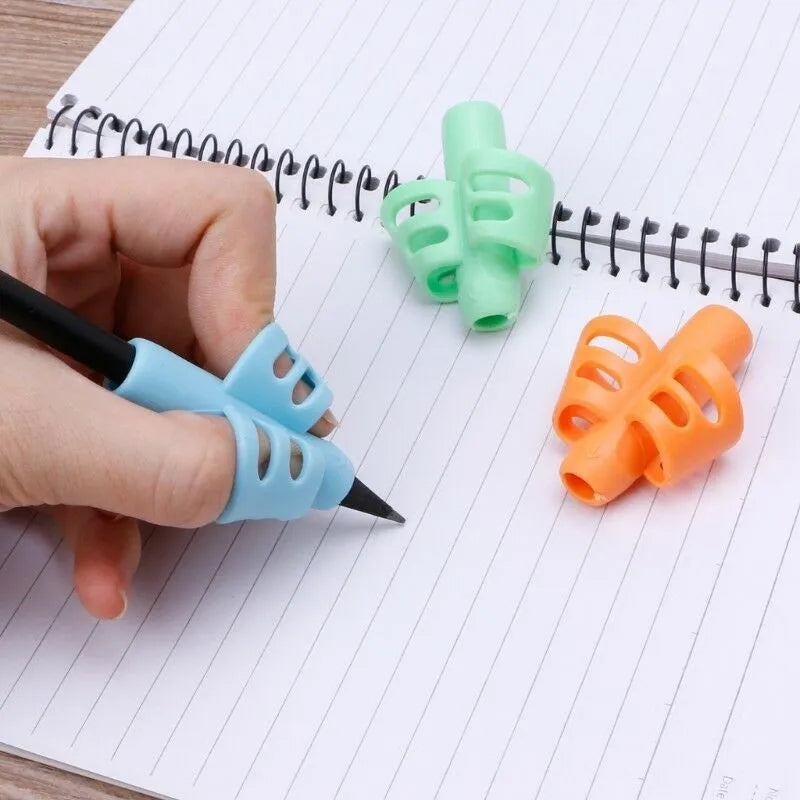 A hand holds a blue silicone pencil grip writing corrector next to green and orange silicone pencil grips on lined paper.