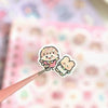 A hand is applying a cute cartoon sticker featuring a smiling, fluffy character to a sheet of patterned paper.