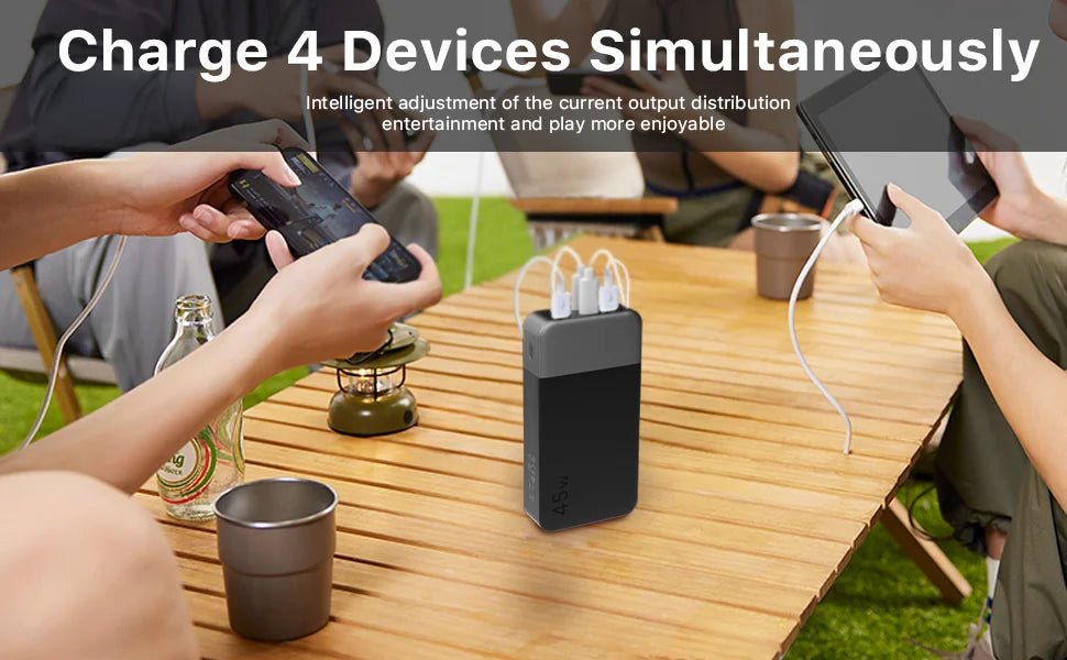 A portable power bank is shown charging four devices simultaneously on a wooden table outdoors.