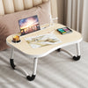 A light wood laptop desk organizer tray with a laptop, paint palette, and a glass sits on a beige upholstered bed.