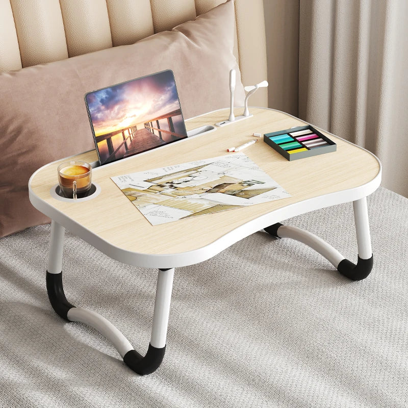 A light wood laptop desk organizer tray with a laptop, paint palette, and a glass sits on a beige upholstered bed.