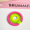 A pink magnetic phone grip and stand with a foldable green ring resting on a white surface.