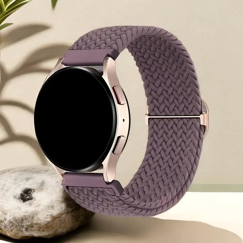 A close-up of a mauve woven nylon watch strap with a rose gold buckle and a black smartwatch face resting on a light surface.