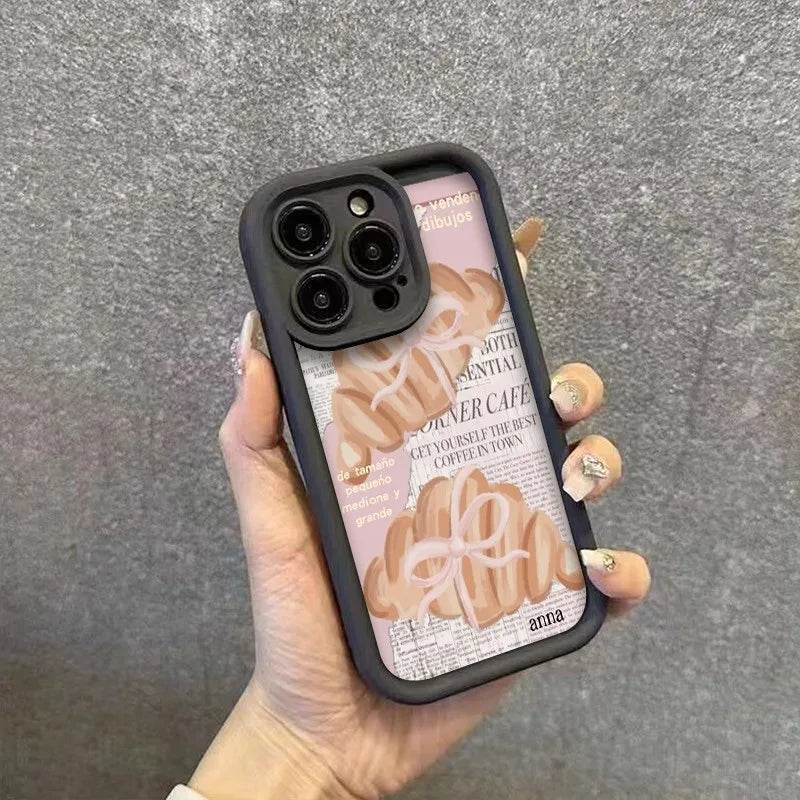 A hand holds a black phone case with a croissant newspaper pattern and a camera lens cover.