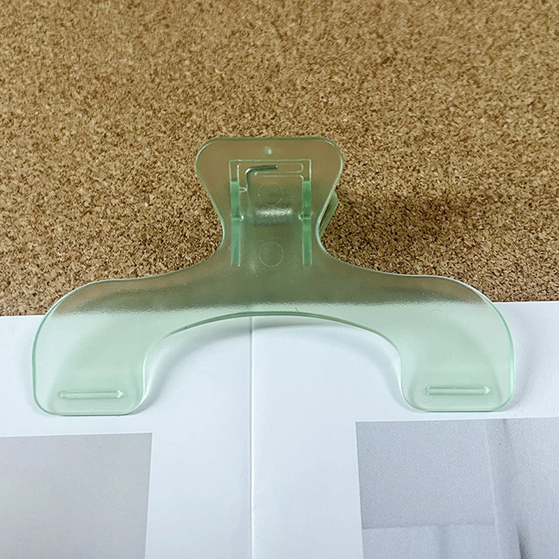 A clear, translucent book page clip is shown resting on a white surface against a textured brown background.
