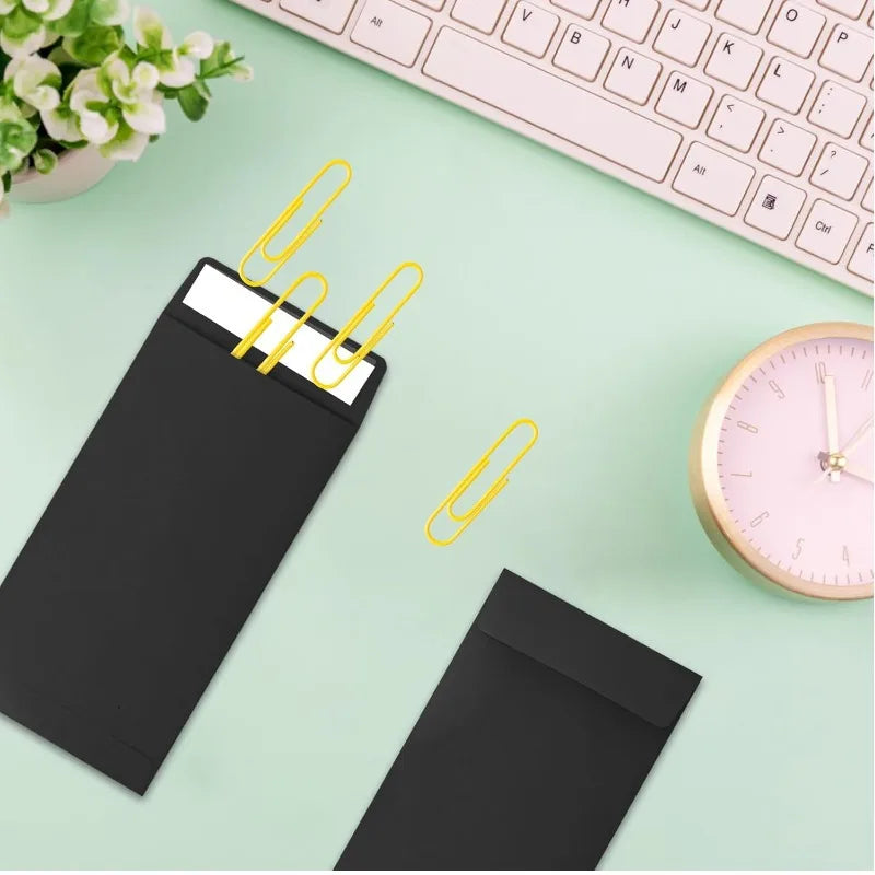 A black kraft envelope is shown with yellow paper clips on a light green surface next to a pink analog watch.
