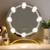 A round LED vanity mirror with multiple bright lights is displayed on a gold stand.