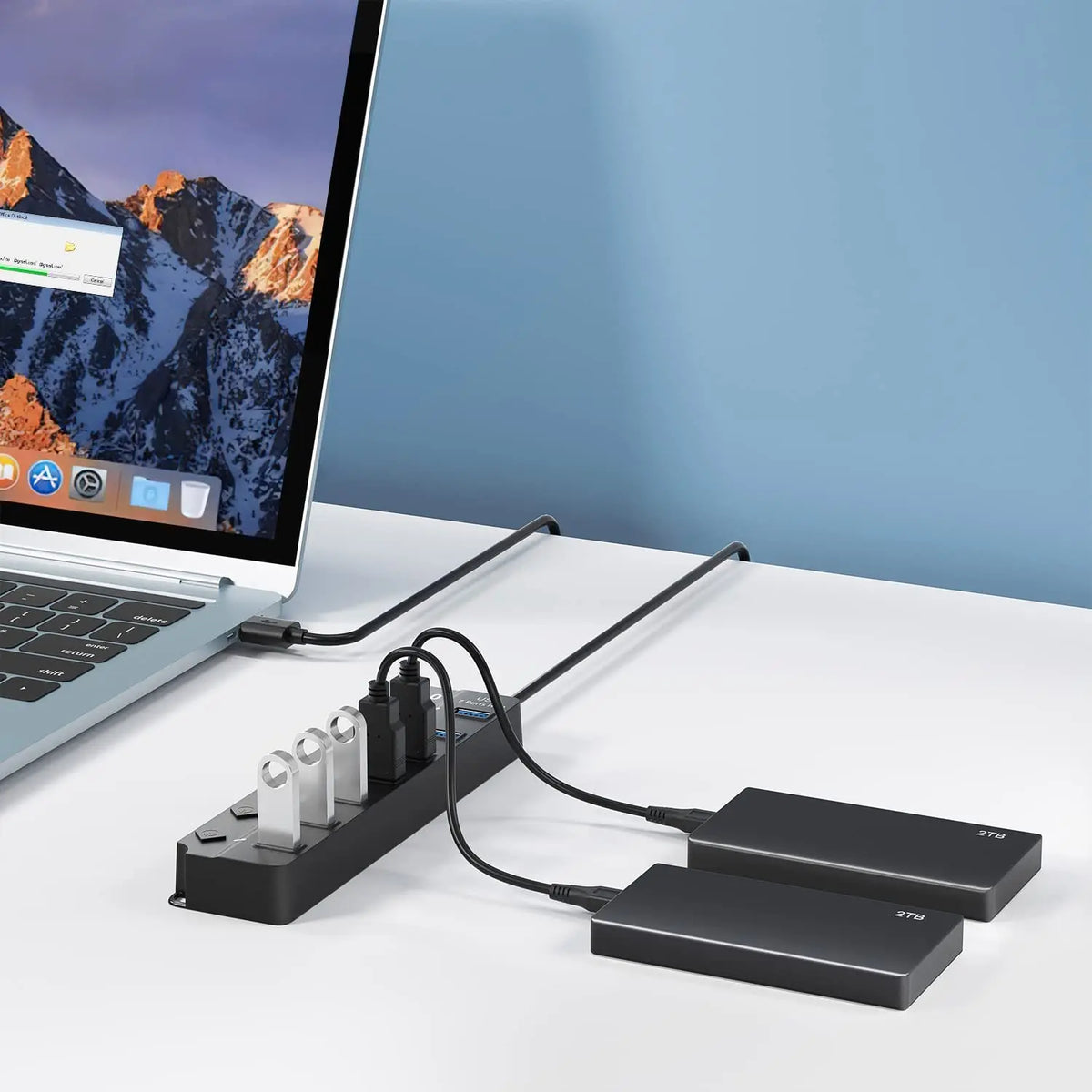 A black USB 3.0 hub extender with multiple cables and power adapters lying on a white desk next to a laptop.