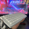 A hand holds a white compact gaming keyboard with RGB backlighting in front of a computer setup.