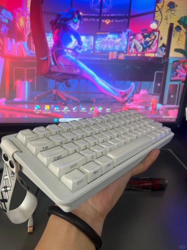 A hand holds a white compact gaming keyboard with RGB backlighting in front of a computer setup.