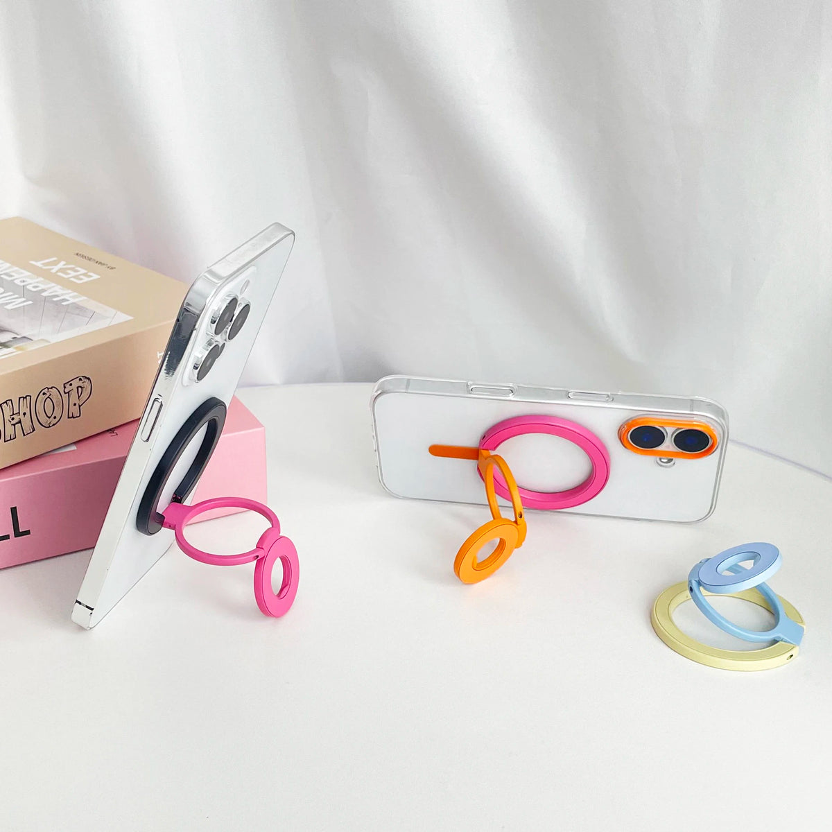 A white smartphone is shown with a pink magnetic grip and an orange foldable ring, next to its packaging box.
