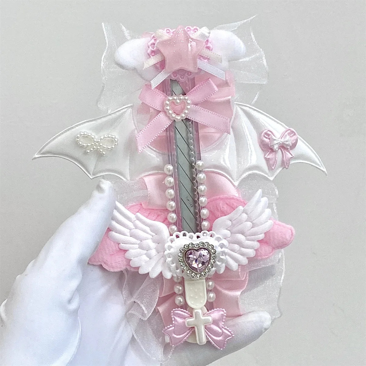 A hand holds an intricate angel wing art knife cutter featuring pink and white fabric details and pearl accents.