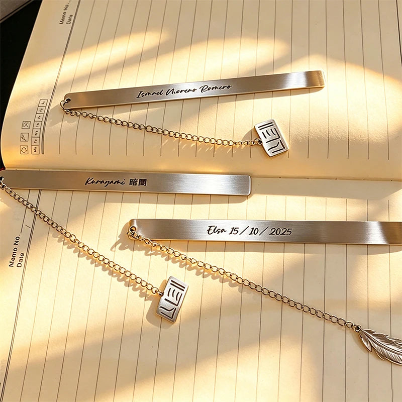 Stainless steel bookmarks with engraved text and a delicate chain, resting on lined paper.