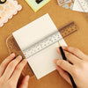Hands are using a black pen to write on a white sheet of paper placed over an acrylic T-square ruler set.