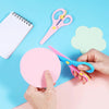 Hands are using pink and blue plastic scissors on a light blue background next to a notebook and a light green paper.