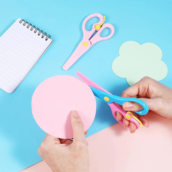 Hands are using pink and blue plastic scissors on a light blue background next to a notebook and a light green paper.