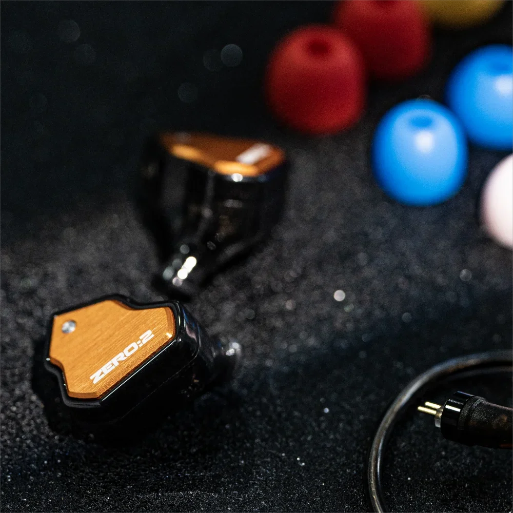 A close-up shot of a black wired in-ear monitor labeled ZERO:2 next to various colored spherical objects.