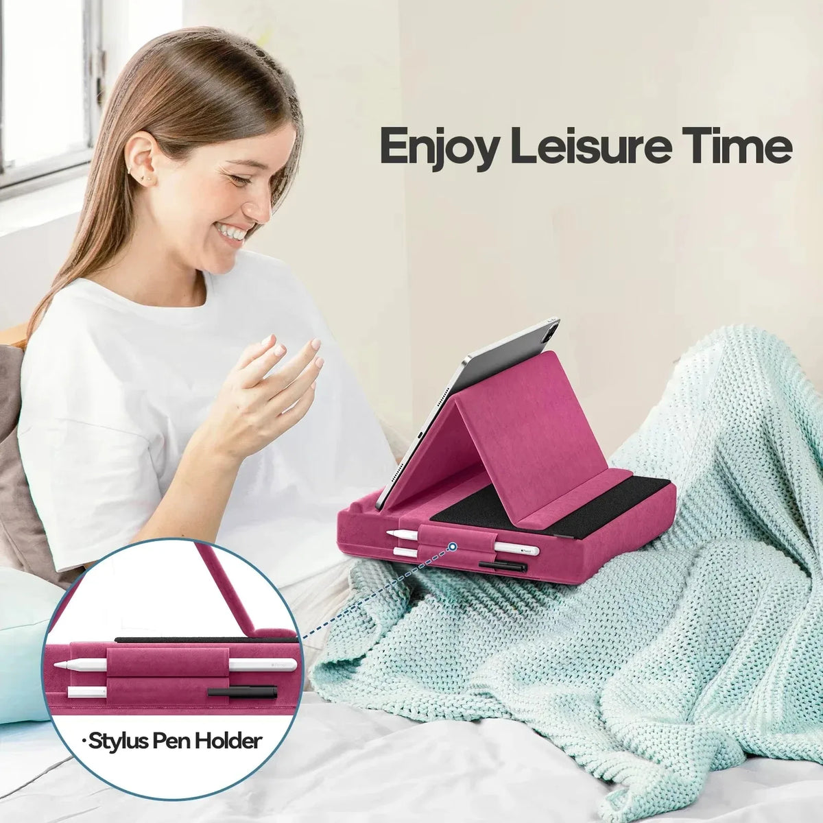 A pink foldable tablet pillow stand with a stylus pen holder is shown resting on a teal blanket next to a smiling woman.