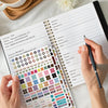 A hand is writing in an undated monthly budget planner with a grid of colored stickers and a notebook.