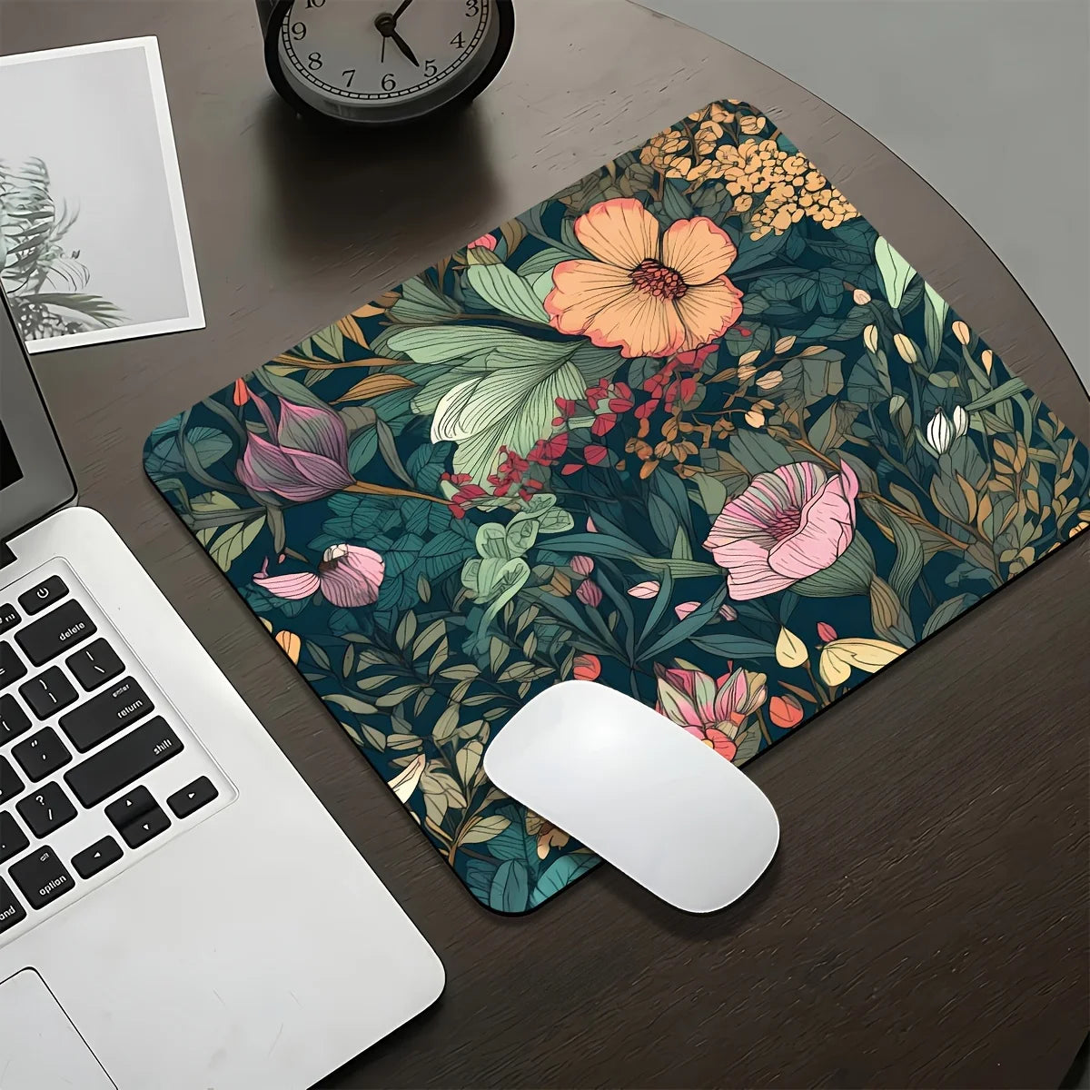A dark floral design mouse pad featuring pink, orange, and green leaves on a wooden desk next to a laptop.