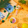 A set of rotating gradient acrylic beaded pens displayed on an orange background with tropical leaves and fruit.