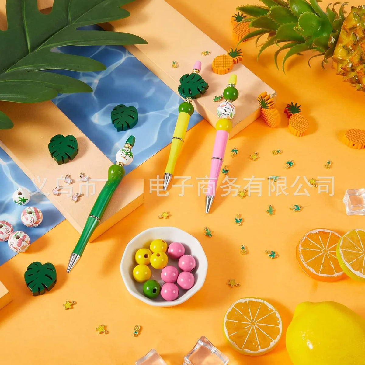 A set of rotating gradient acrylic beaded pens displayed on an orange background with tropical leaves and fruit.