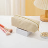 A beige zippered pencil case resting on a white surface next to makeup brushes and a small electronic device.