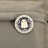 An enamel pin featuring a white ghost with butterfly wings and floral accents on a black background.