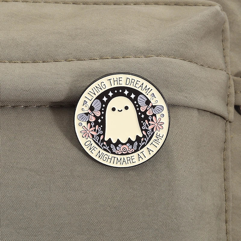 An enamel pin featuring a white ghost with butterfly wings and floral accents on a black background.