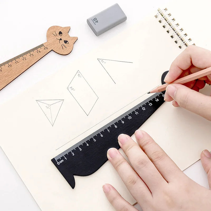 A hand draws geometric shapes on paper using a wooden ruler and pencil next to a cat-shaped drawing tool.