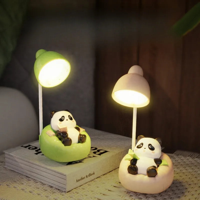 A white panda-shaped LED desk lamp with a mobile phone holder sits on a book next to a green lamp.