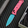 A pink acrylic folding knife with a D2 steel blade is shown resting on a dark surface.