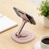 A pink foldable desktop phone stand holding a smartphone, placed on a wooden table next to a notebook and a cup.