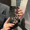 A hand holds a black phone case featuring gothic floral art on an iPhone against a gray background.