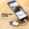 A hand holds a smartphone displaying a photo, next to a USB-C SD card reader device.