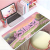 A large pink and green The Sims gaming mouse pad is displayed on a white desk next to a laptop and stationery.